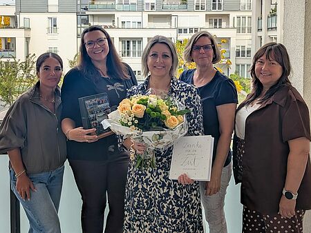 Team Pflegedienstleitung Das Team Pflegedienstleitung steht auf dem Balkon als Gruppe zusammen, zur Verabschiedung hat eine der Kolleginnen einen Blumenstrauß und Geschenke überreicht bekommen, die sie nun in Händen hält